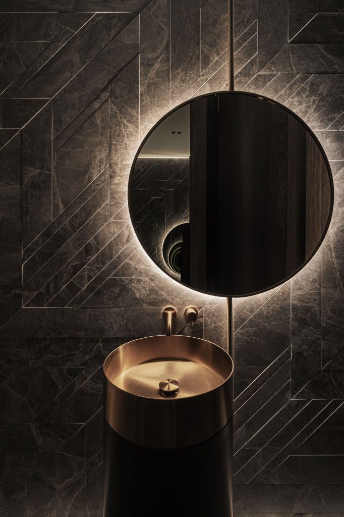 Dark grey geometric marble walls with cylindrical brass sink and back-lit circle mirror by YODEZEEN studio.